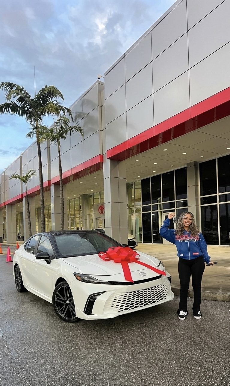 Customer holding up keys beside a white sedan with a red bow outside a dealership.